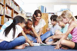 A group of children sit on the floor of a library while an adult points to something in a picture book.