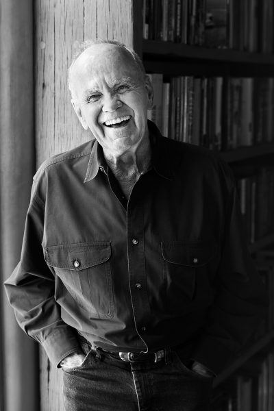 Black and white photo of Cormac McCarthy standing in front of a bookshelf wearing a dark button-up shirt and dark jeans, and a belt with a silver and turquoise Native American style buckle. His hands are in his pockets and he is laughing.