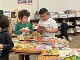 Two kindergarten students look though books laid out on a table.