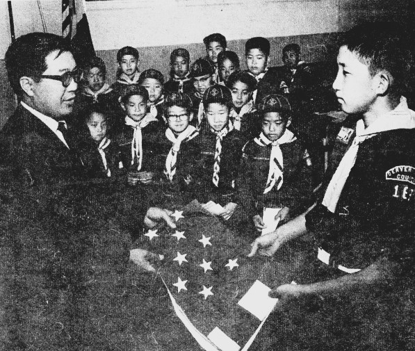 A boy scout troops looks on as one of their members receives a U.S. flag from an official.