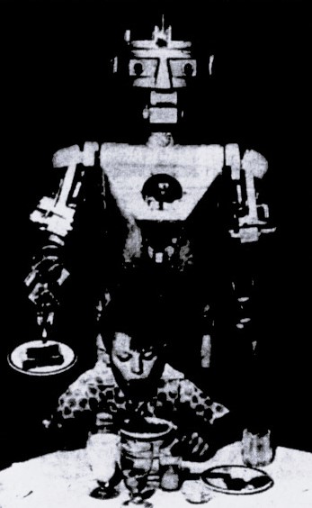 A robot stands behind a boy at a table and serves him food.
