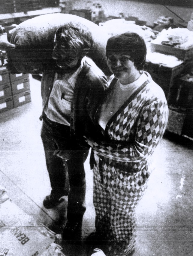 A woman in an argyle outfit stands next to a man carrying a sack of wheat.