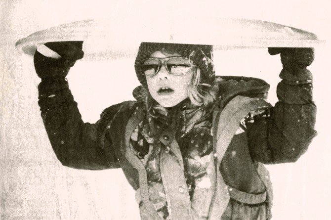 A girl wearing winter clothes and sunglasses carries a snow saucer above her head.