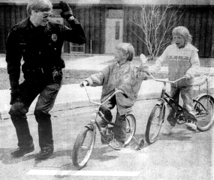 A police officer demonstrates how to signal a right turn to two young biker riders.