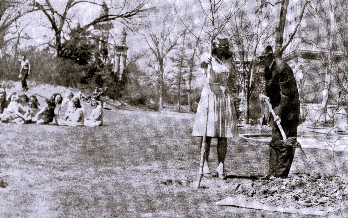 A high school principal shovels dirt around a tree that a young woman holds.