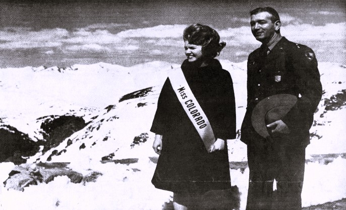 A woman wearing a pageant sash and a man in a military uniform view a mountain landscape.