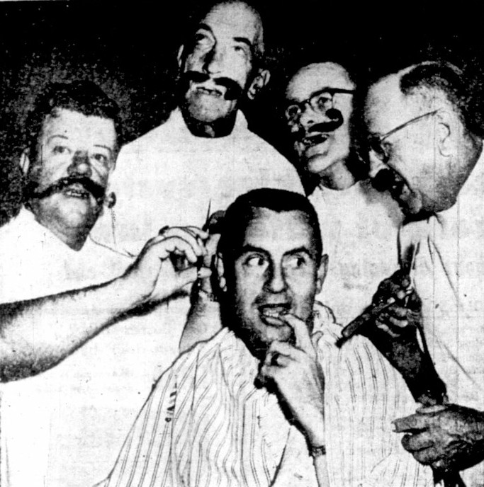 4 men in a barbershop quartet sing to a man in a barber cape.