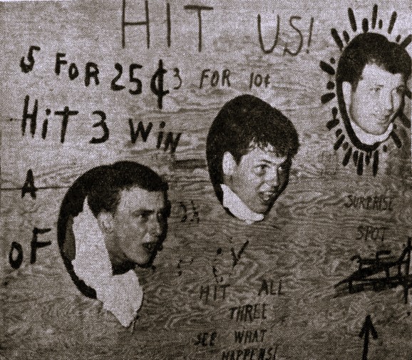 Three young men stick their heads through a piece of wood at a carnival booth.