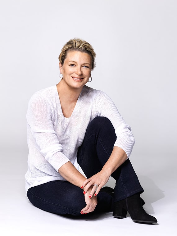 Portrait of Kristin Hannah. She is sitting on the floor holding her bent left knee. Wearing black trousers and boots and a whit V-neck sweater. Her hair is pulled back in a ponytail and she is wearing hooped earrings. She is smiling.