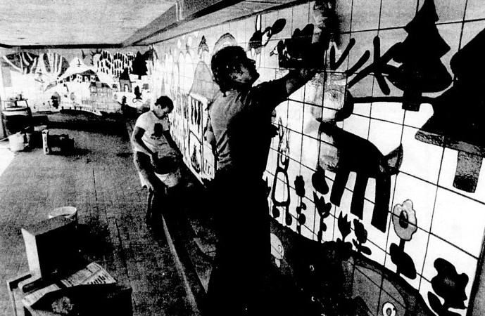 Two men paint the walls of a bus stop with designs created by children.