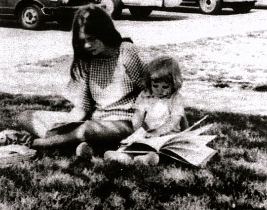 An older and younger girl read books on the grass.