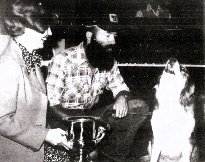 A woman awards a trophy to a man and his howling dog.