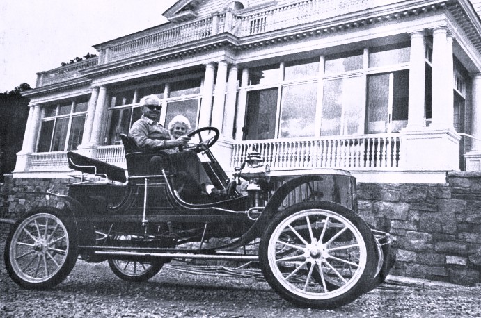 A man and woman sit in an early model automobile.