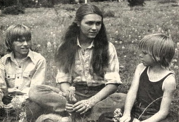 A young woman and two boys sit on the grass holding flowers.