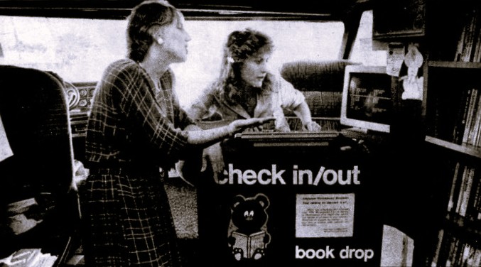 A librarian demonstrates a computer checkout system to another woman.