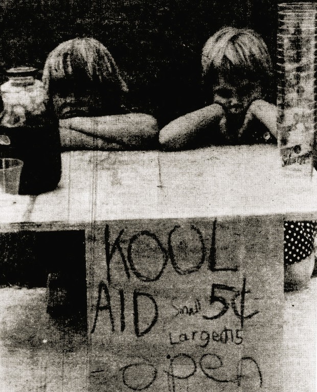 Two children sit at a Kool-Aid stand.