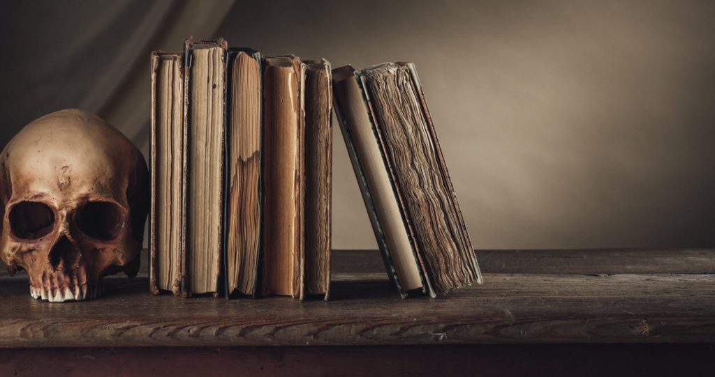 A dark image of a human skull serving as a book end for a row of seven very old looking books. The spines are facing away so that the titles cannot be read. They are on a wood shelf.