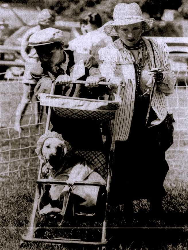 A woman and a boy push a dog wearing a bonnet in a stroller.