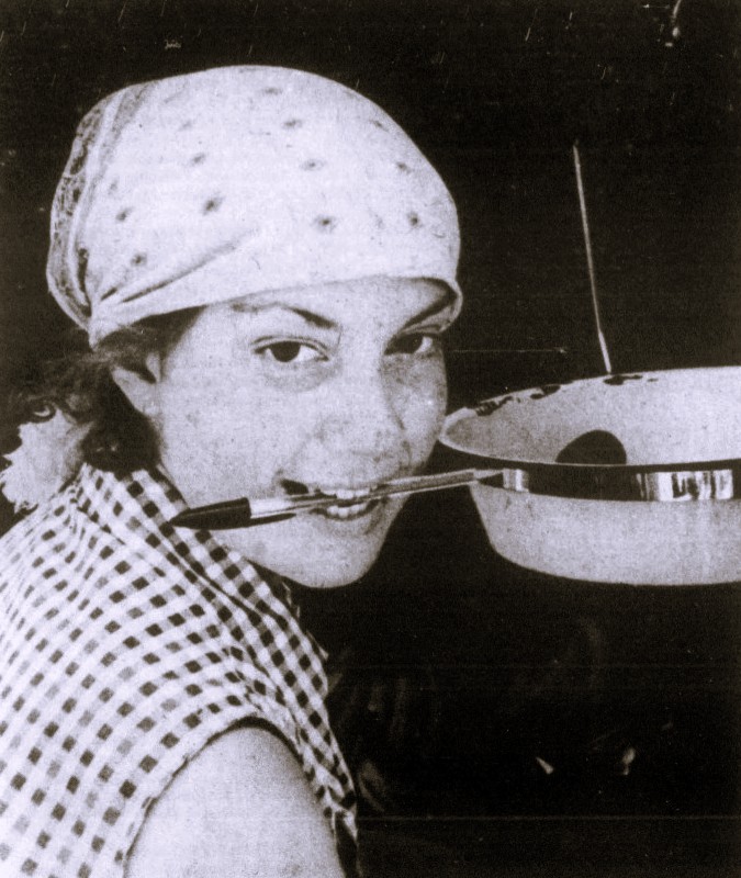 A woman wearing a bandana holds a pen in her mouth.