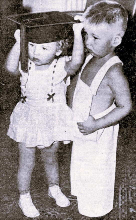 A boy toddler in overalls stands next to a girl toddler wearing a graduation cap.