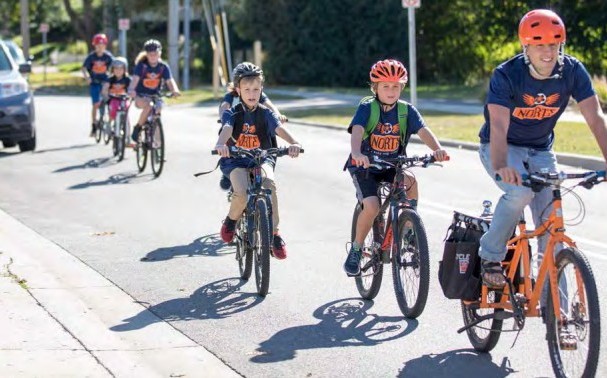 A teacher leads a line of school-age kids riding bikes, all wearing matching t-shirts with their school logo.