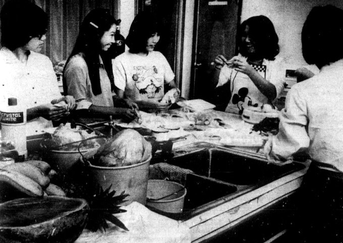 A woman wearing an apron demonstrates a cooking technique to 4 students.