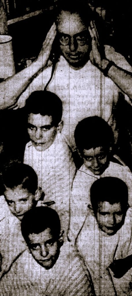 A barber stands behind 5 brothers awaiting haircuts.