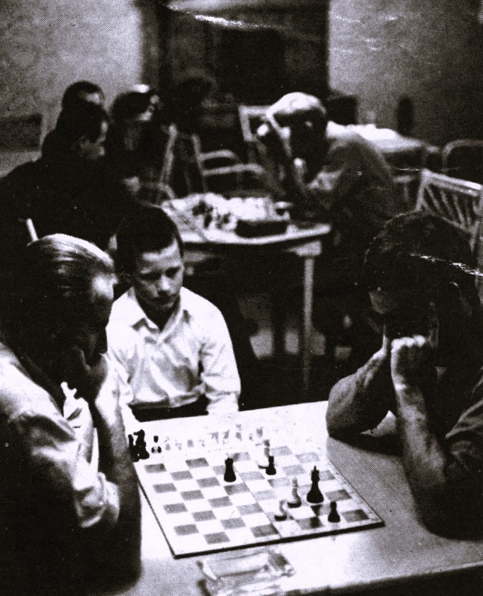 A boy watches two men competing in a chess tournament.