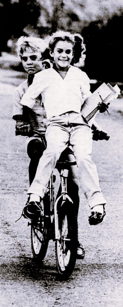 A boy rides a bike with a girl on the handlebars.