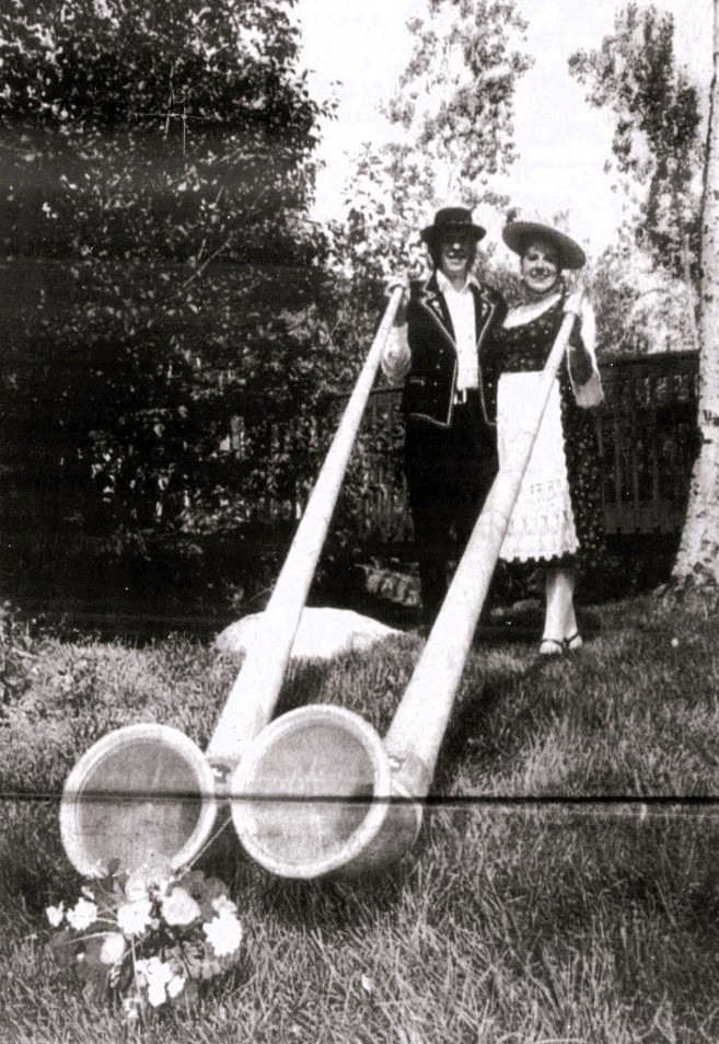 Two alphorn musicians in traditional dress stand with their instruments.