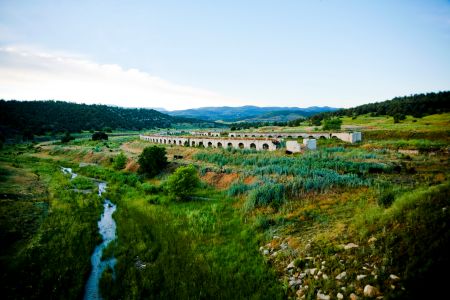 A double line of large, white industrial ovens made of stone, with large half-circular mouths, sit next to a stream in a mountain landscape.