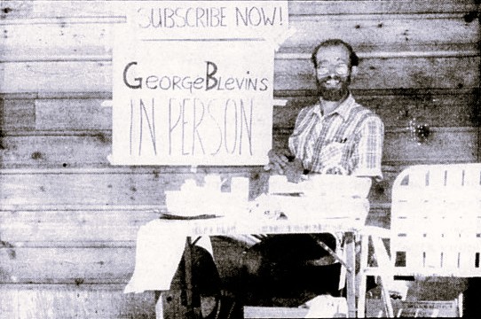 A man sits at a table with a sign reading 'George Blevins In Person'.