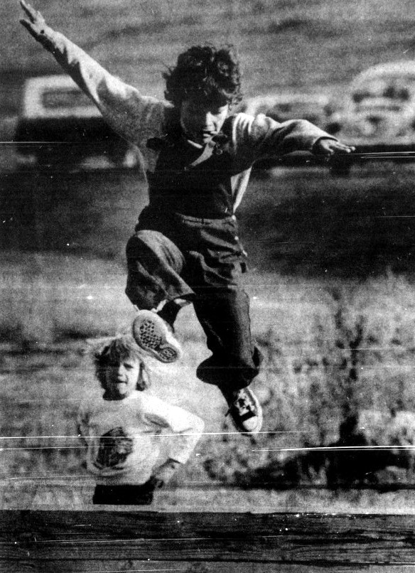 A boy jumps over an obstacle while another boy watches.