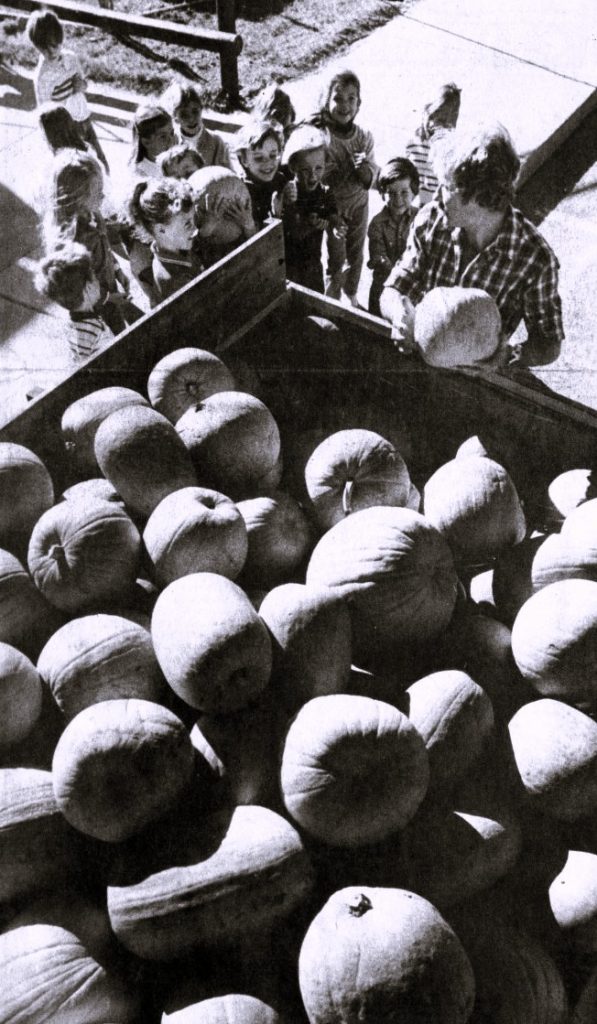 A group of children gather around a man handing out pumpkins from a truck bed.