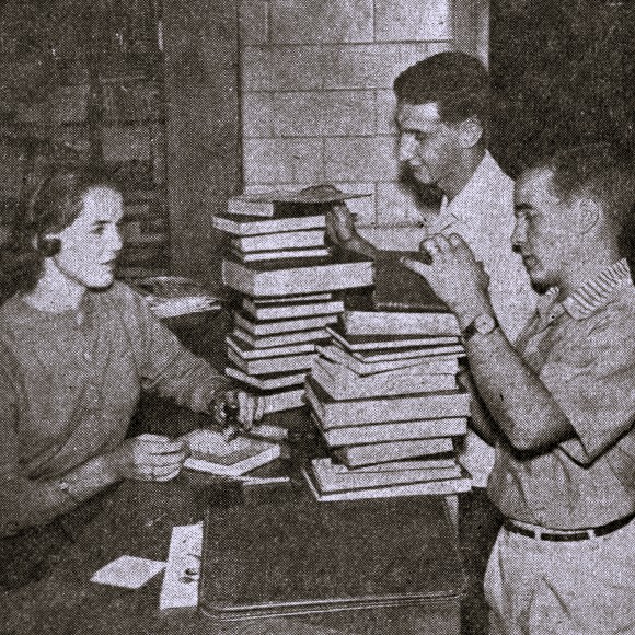 A young woman at a library circulation desk checks out stacks of books to two young men.