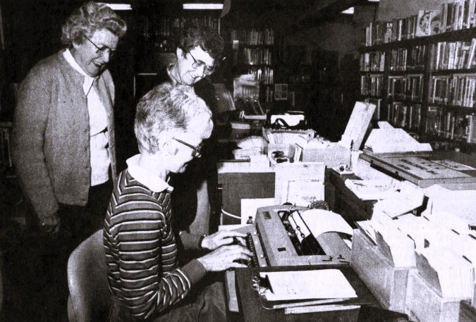 Three women in a library admire a new typewriter.