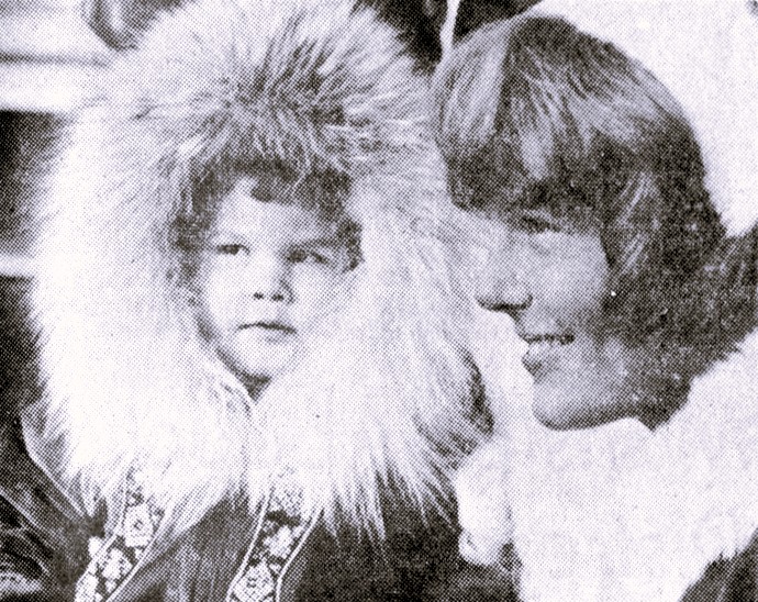 A girl in a fur-lined parka stands beside a woman in a Christmas costume.