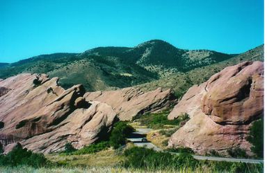 A two-lane, paved road winds through a rocky landscape, with green hills in the background.