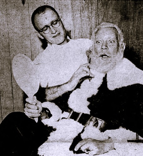 A barber trims Santa's beard while he looks in a hand mirror.
