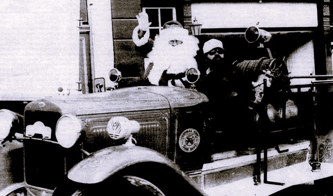 Santa waves from a vintage firetruck driven by a man in hat and sunglasses.