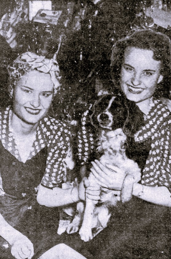 Twin sisters in matching outfits hold a small dog.