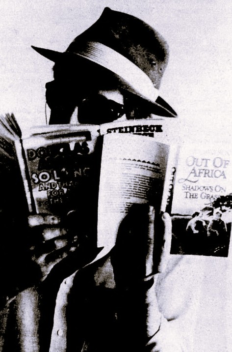 A man in a fedora hat and sunglasses holds up three books.