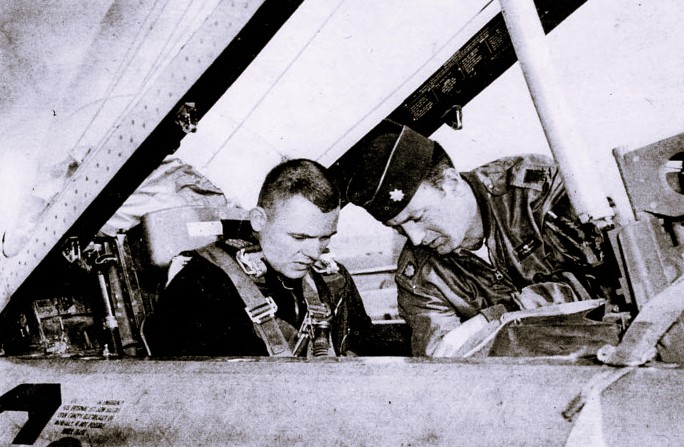 An airman sits in a jet cockpit while his instructor leans in.