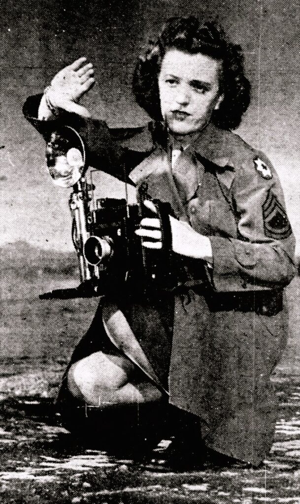 A woman in military dress crouches while holding a camera.