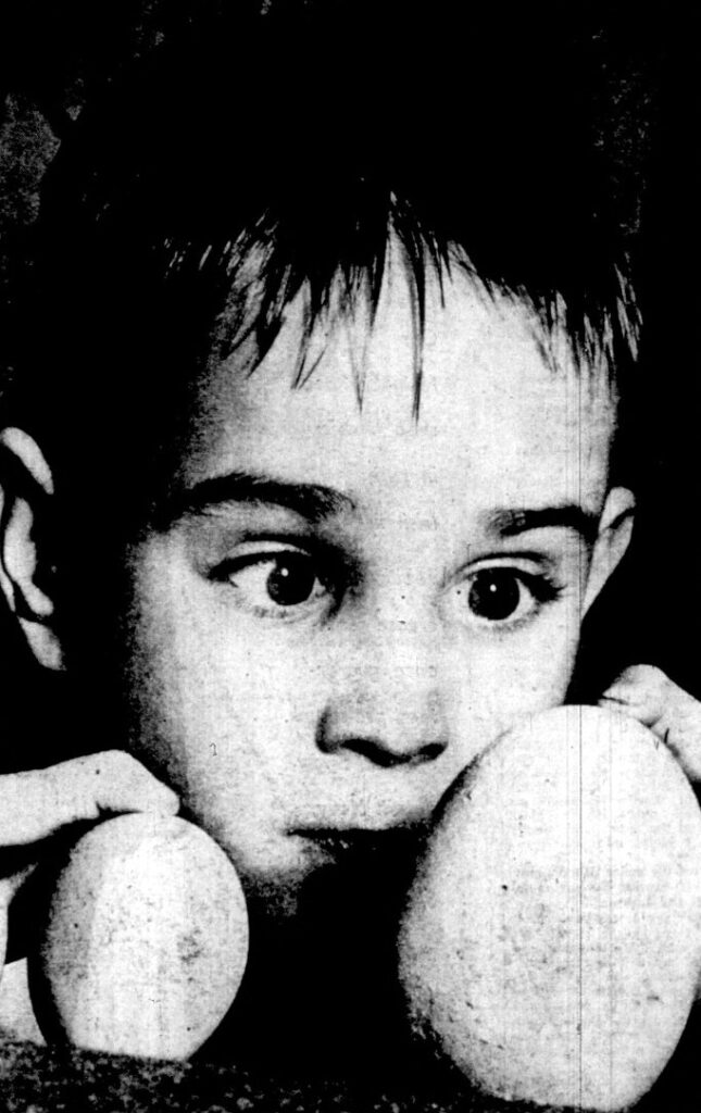 A young boy stares at a very large chicken egg.