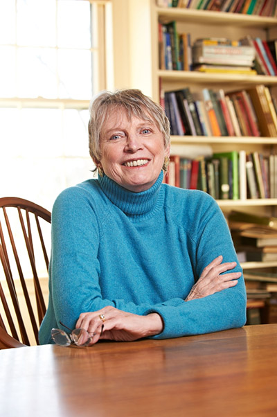 Lois Lowry sitting at a wooden table wearing a turquois turtleneck sweater. She is in front of a bookcase to the right of the image.
