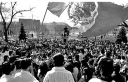A large group of people listening to a speaker at the Colorado capitol building stand underneath a Mexican flag.