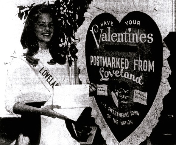 A young woman wearing a sash stands next to a large heart.