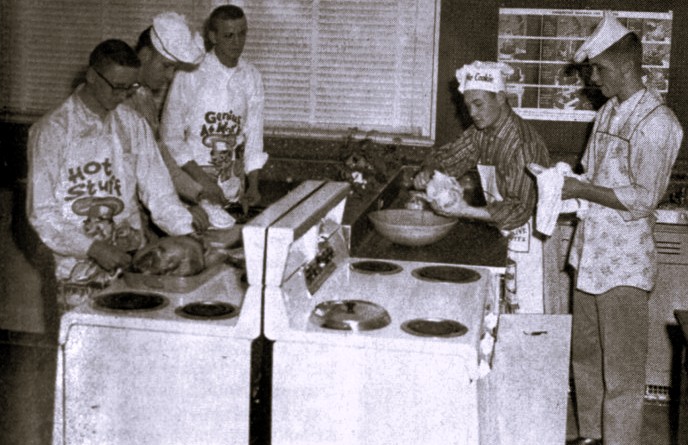 Five high school boys wearing aprons prepare a meal in a classroom kitchen.