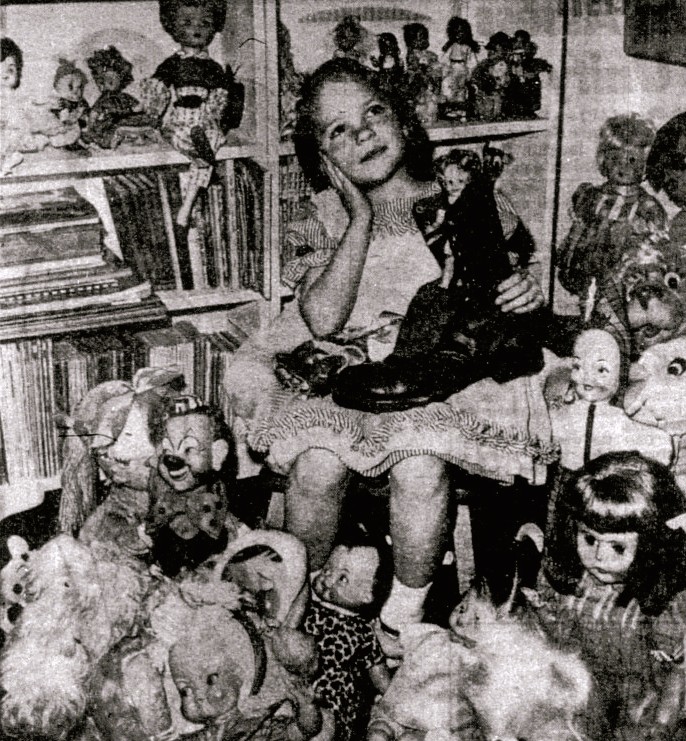 A girl sits surrounded by dolls.